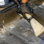 A hand in a glove using a vacuum to clean a car interior carpet, removing dirt effectively.